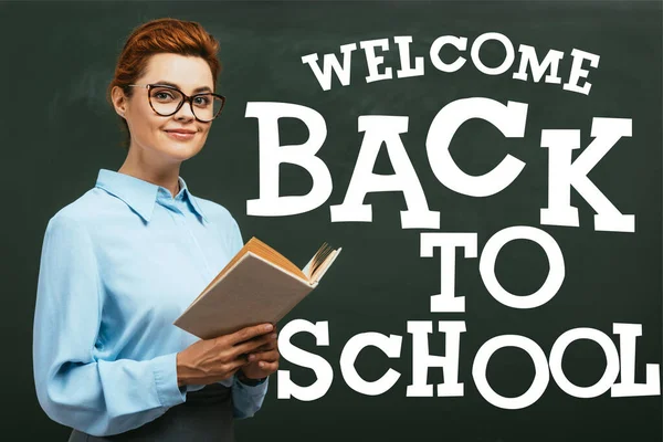 Teacher in eyeglasses holding book near chalkboard with welcome back to school lettering in classroom — Stock Photo