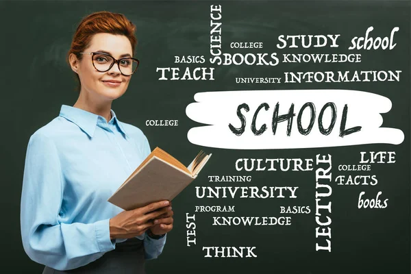 Teacher in eyeglasses holding book near chalkboard with lettering in classroom — Stock Photo