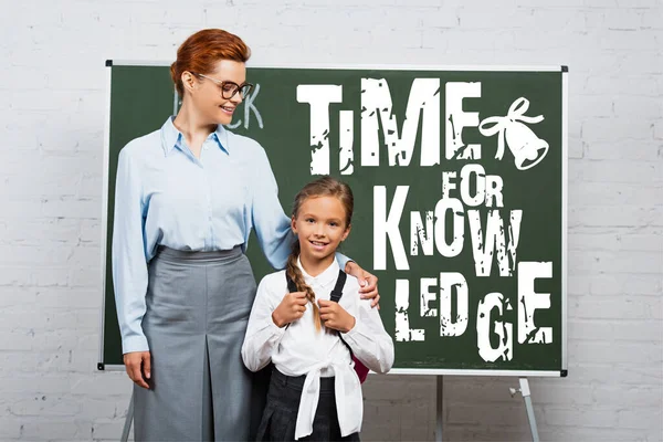 Teacher and schoolgirl standing near chalkboard with time for knowledge lettering — Stock Photo