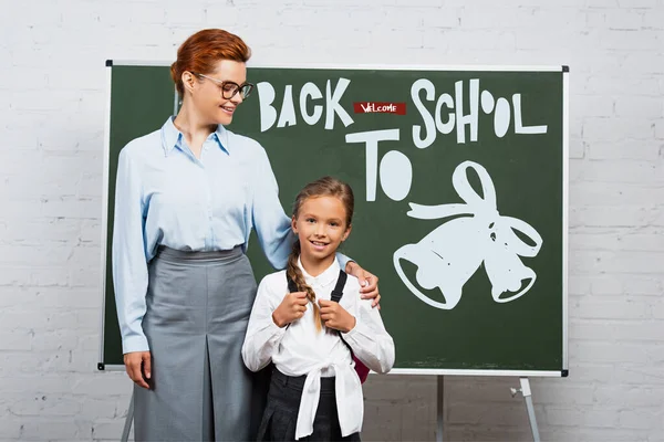 Teacher and schoolgirl standing near chalkboard with back to school lettering — Stock Photo