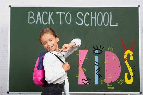 Schoolgirl pointing with finger at backpack near chalkboard with back to school and kids lettering in classroom — Stock Photo