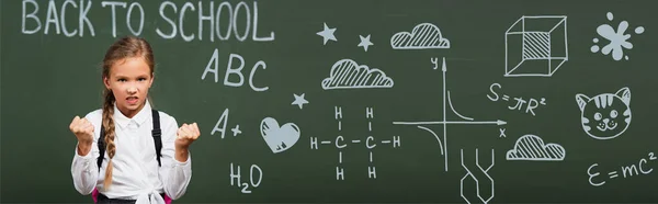 Horizontal image of angry schoolgirl showing clenched fists near chalkboard with back to school lettering and geometry formulas in classroom — Stock Photo