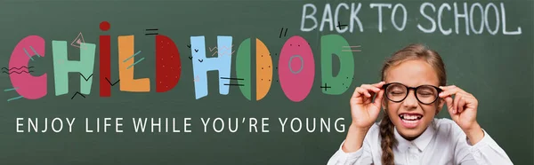 Horizontales Bild von Schulmädchen berühren Brillen in der Nähe der Tafel mit dem Rücken zur Schule und Kindheit, genießen Sie das Leben, während Sie jung sind Schriftzug — Stockfoto