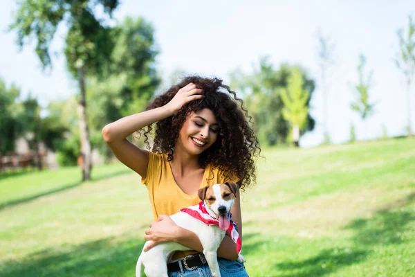 Mulher alegre tocando cabelo encaracolado enquanto segurando Jack Russel cão terrier no parque — Fotografia de Stock