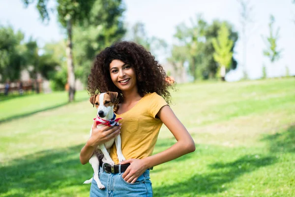 Young woman in summer outfit standing with hand in pocket while holding jack russell terrier dog in park — Stock Photo