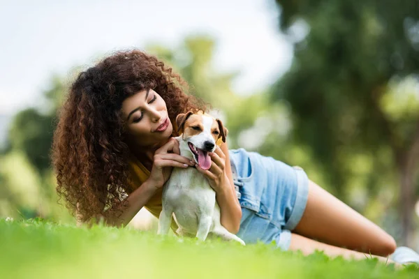 Vista livello della superficie della giovane donna riccia abbracciando jack russell terrier cane mentre si trova sul prato verde — Foto stock
