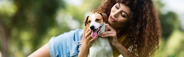 Horizontal orientation of curly woman cuddling jack russell terrier dog while resting in park — Stock Photo