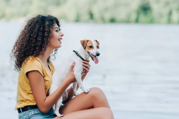 Side view of excited woman in summer outfit sitting near lake with jack russell terrier dog — Stock Photo