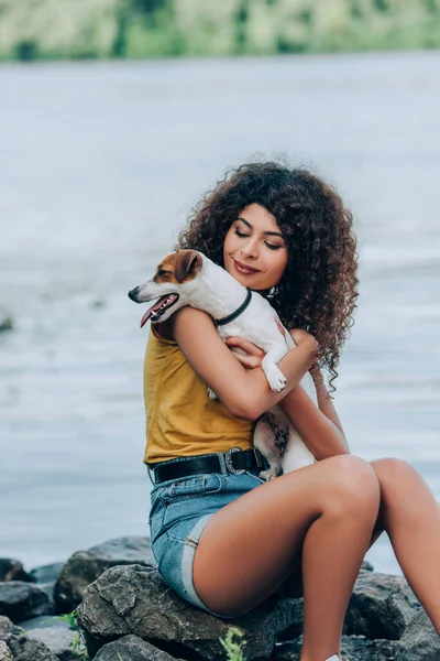 Morena, rizado mujer en verano traje celebración jack russell terrier perro mientras sentado en piedras cerca de río - foto de stock