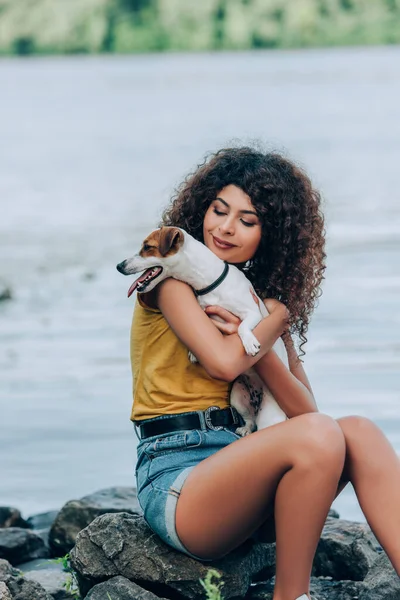 Morena, mulher encaracolada em roupa de verão segurando jack russell terrier cão enquanto sentado em pedras perto do rio — Fotografia de Stock