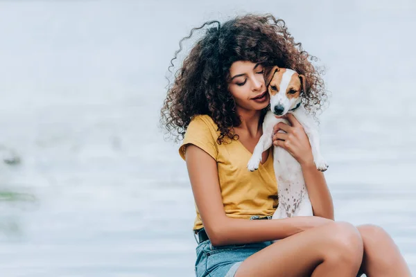 Rizado mujer en verano traje abrazando jack russell terrier perro cerca de lago - foto de stock