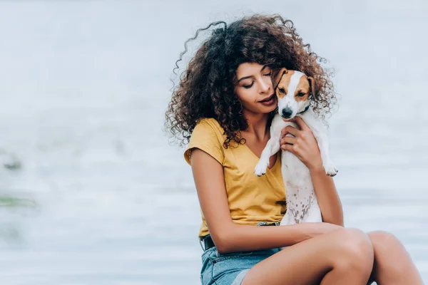 Mulher encaracolado em roupa de verão abraçando Jack Russell cão terrier perto do lago — Fotografia de Stock