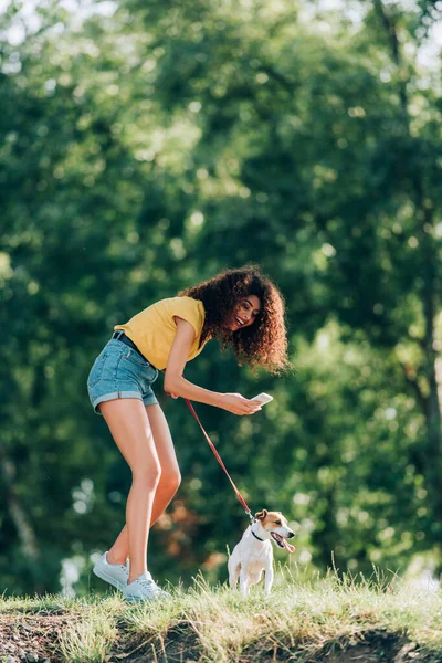 Young woman in summer outfit laughing while taking photo of jack russell terrier dog in park — Stock Photo