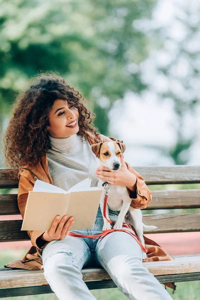 Femme élégante regardant loin tout en tenant le livre et câlin Jack Russell Terrier chien sur le banc dans le parc — Photo de stock
