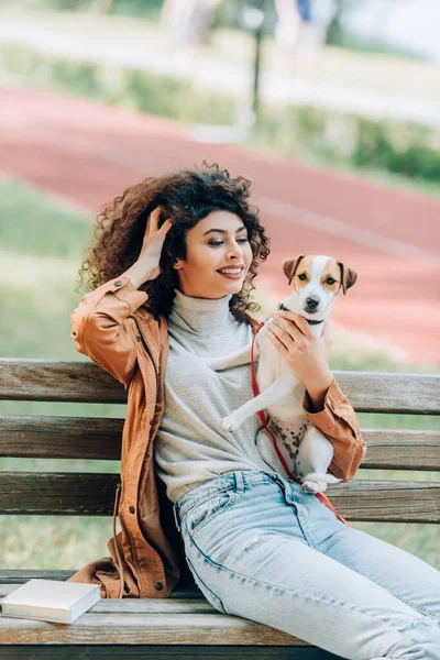 Joyful woman in autumn outfit touching curly hair while sitting with jack russell terrier dog on bench in park — Stock Photo
