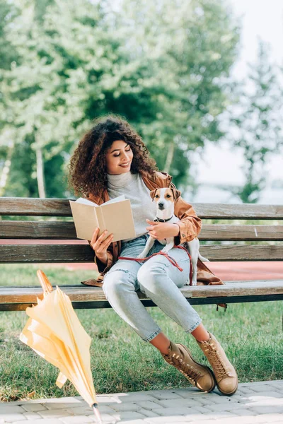 Femme élégante en tenue d'automne tenant livre et câlin Jack Russell Terrier chien sur banc dans le parc — Photo de stock