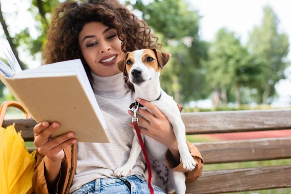 Joyful woman reading book and holding jack russell terrier dog on bench in park — Stock Photo