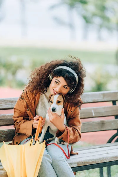 Mulher encaracolado em fones de ouvido sem fio abraçando Jack Russell cão terrier enquanto sentado no banco com guarda-chuva amarelo — Fotografia de Stock