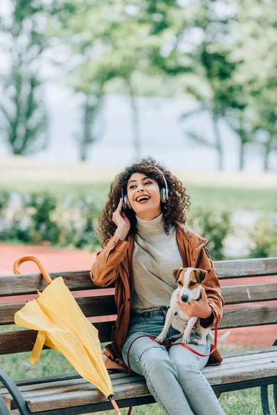Mujer riendo en auriculares inalámbricos mirando hacia otro lado mientras está sentado en el parque con Jack Russell terrier perro cerca de paraguas amarillo - foto de stock