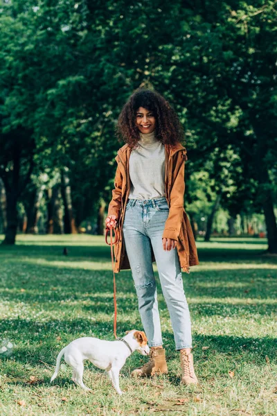 Selektiver Fokus einer Frau im Regenmantel, die in der Nähe von Jack Russell Terrier im Park in die Kamera blickt — Stockfoto