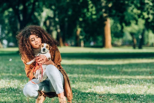 Donna riccia in impermeabile abbracciando jack russell terrier sul prato nel parco — Foto stock