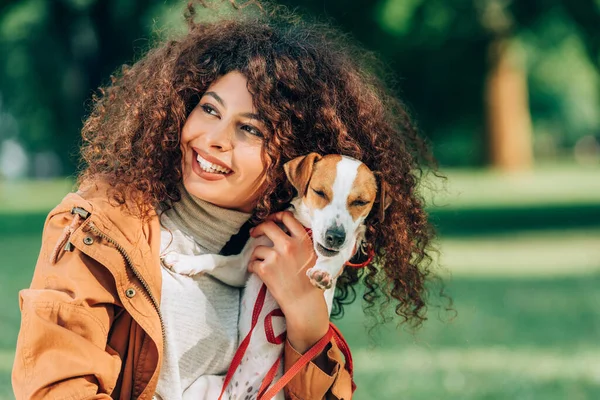Selective focus of curly woman in raincoat looking away while holding jack russell terrier in park — Stock Photo