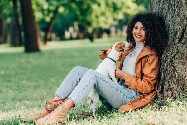 Selektiver Fokus einer jungen Frau im Regenmantel, die mit Jack Russell Terrier spielt und in der Nähe eines Baumes im Park in die Kamera blickt — Stockfoto