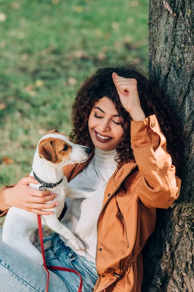 Curly woman in raincoat holding jack russell terrier near tree in park — Stock Photo