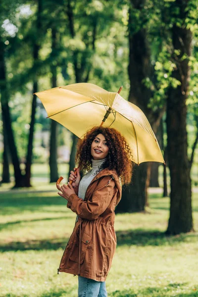 Кудрявая женщина в плаще держит зонтик и смотрит в камеру в парке — стоковое фото