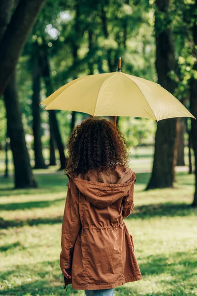 Vue arrière de la femme en imperméable tenant parapluie jaune dans le parc — Photo de stock