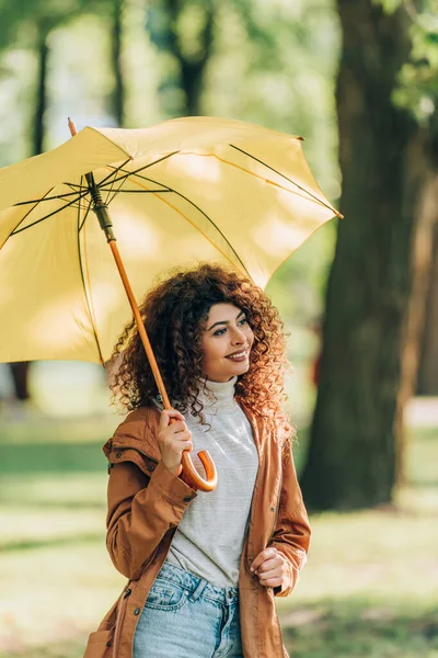 Curly woman in raincoat looking away while holding umbrella in park — Stock Photo