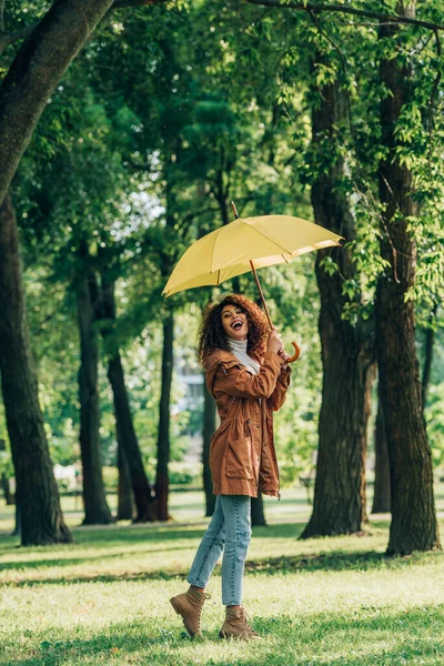 Молодая женщина в осеннем наряде держит желтый зонтик, стоя на траве в парке — стоковое фото