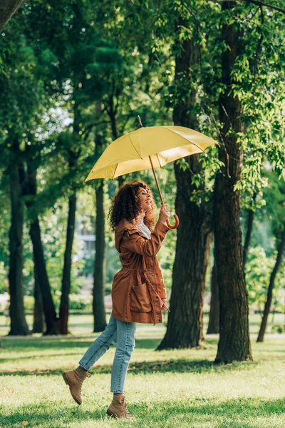 Вид сбоку на кудрявую женщину в плаще и джинсах, держащую зонтик в парке — стоковое фото