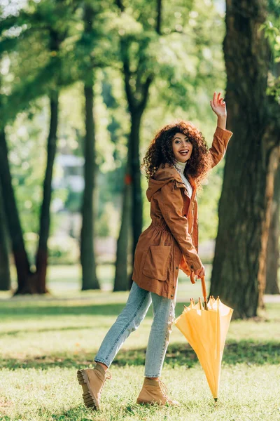 Vue latérale de la femme bouclée avec parapluie agitant la main à la caméra tout en marchant dans le parc — Photo de stock