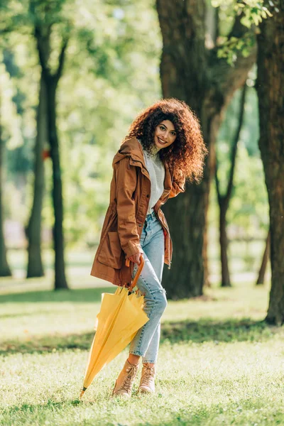 Selektiver Fokus einer lockigen Frau mit gelbem Regenschirm auf dem Rasen im Park — Stockfoto