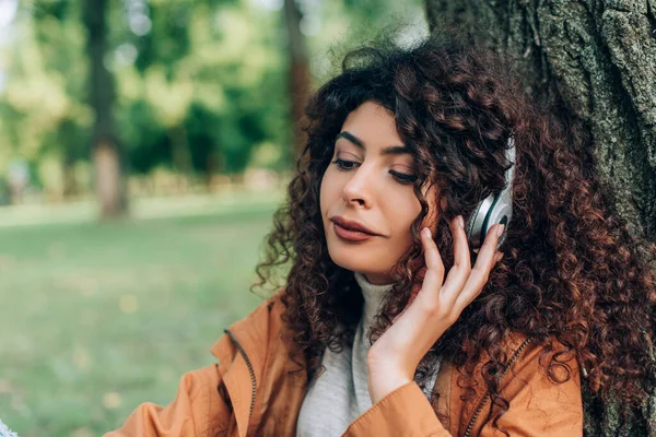 Curly woman listening music in headphones near tree in park — Stock Photo