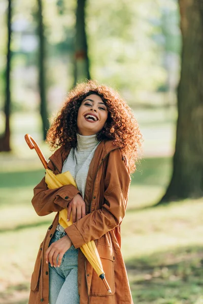 Excited woman in raincoat looking at camera while holding umbrella in park — Stock Photo