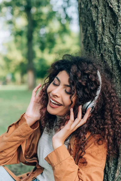 Mujer joven con los ojos cerrados escuchando música en los auriculares cerca del árbol en el parque - foto de stock