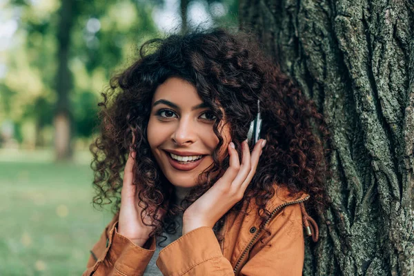 Junge Frau berührt Kopfhörer und blickt in Park in Kamera — Stockfoto