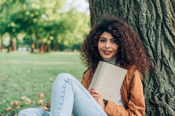 Вибірковий фокус кучерявої жінки в плащі, що тримає книгу біля дерева в парку — стокове фото