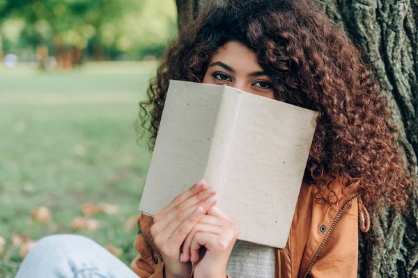 Focus selettivo della donna riccia che tiene libro vicino al viso nel parco — Foto stock