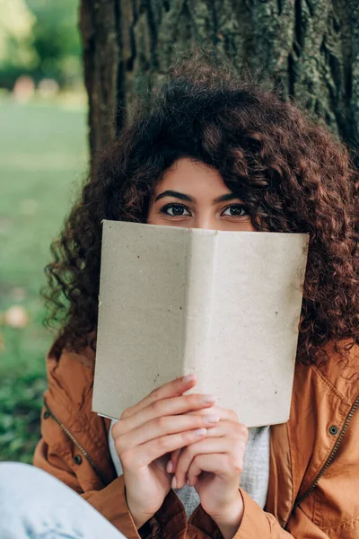 Concentration sélective de la femme regardant la caméra tout en tenant le livre dans le parc — Photo de stock