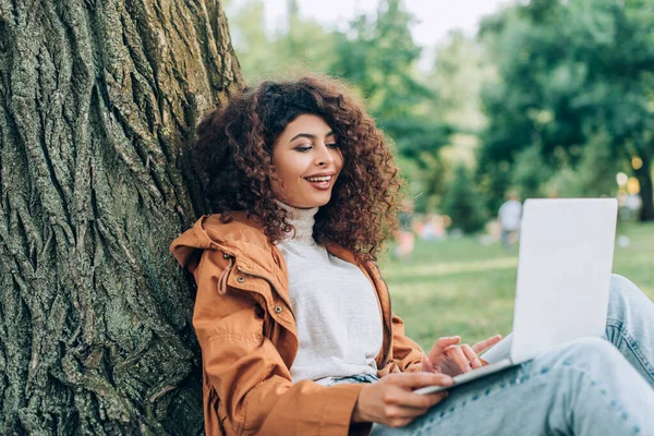 Selective focus of freelancer using laptop near tree in park — Stock Photo
