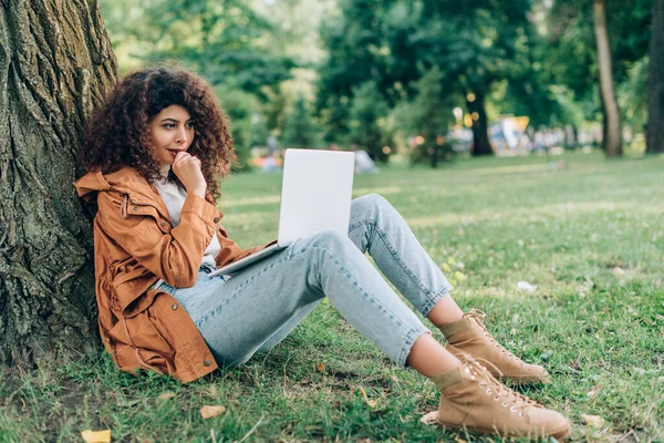 Selective focus of thoughtful woman in raincoat using laptop on grass near tree in park — Stock Photo