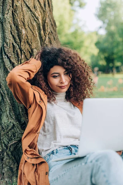 Enfoque selectivo del freelancer en traje de otoño usando portátil cerca del árbol en el parque - foto de stock