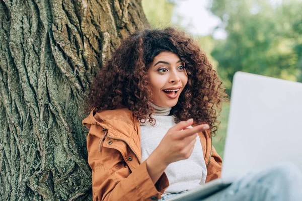 Selective focus of excited freelancer pointing with finger at laptop near tree in park — Stock Photo