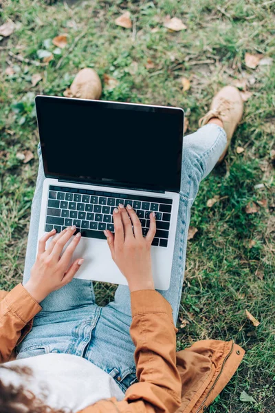 Top view of young woman in raincoat and jeans using laptop with blank screen in park — Stock Photo