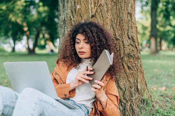 Concentration sélective de la femme regardant l'ordinateur portable tout en tenant un cahier et un stylo près de l'arbre dans le parc — Photo de stock