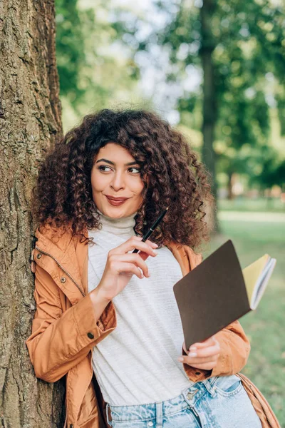 Enfoque selectivo de la mujer rizada en impermeable mirando hacia otro lado mientras sostiene la pluma y el cuaderno en el parque - foto de stock