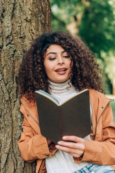 Selective focus of brunette woman in raincoat looking at notebook beside tree in park — Stock Photo